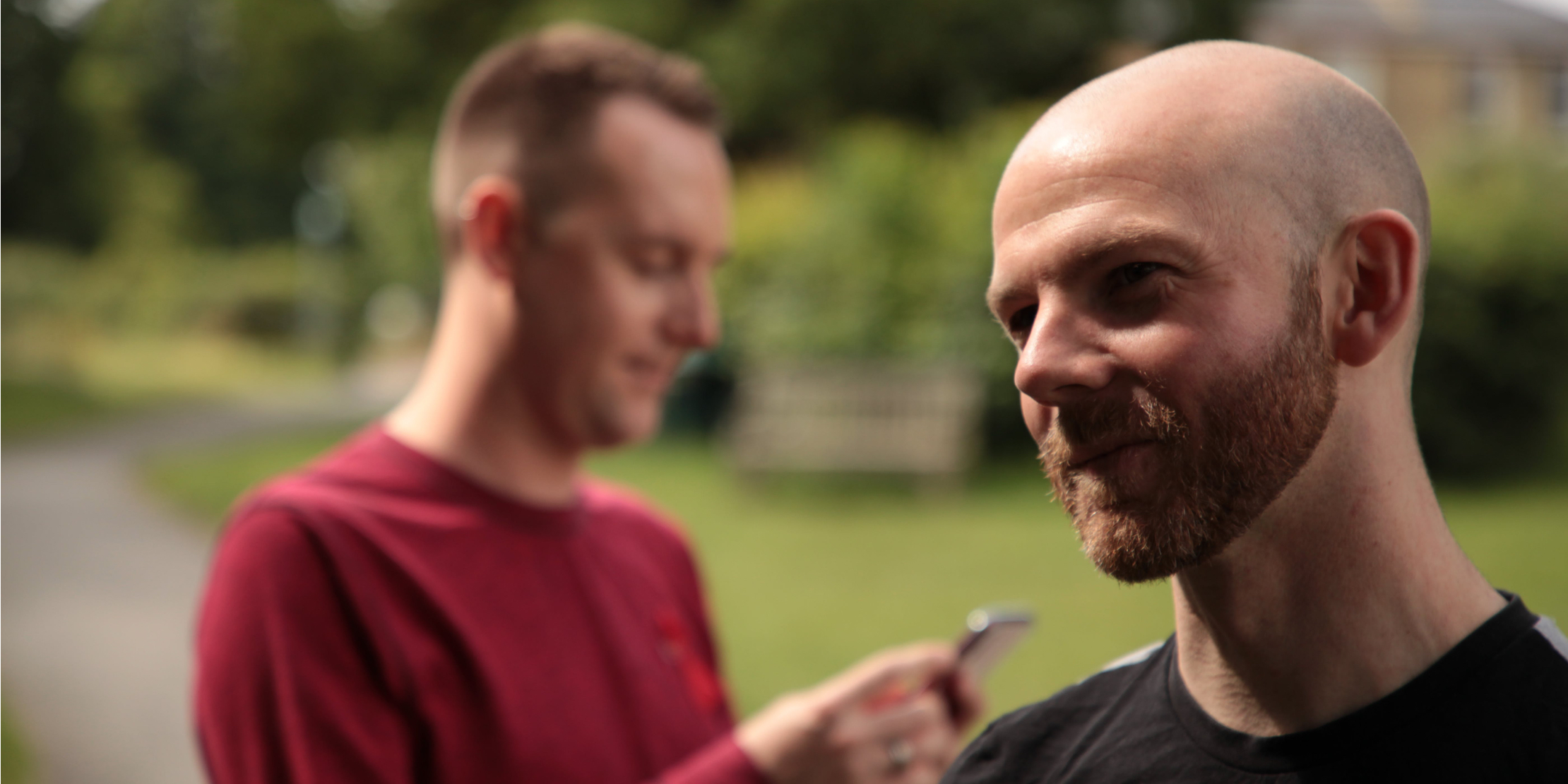 Two men outside; one (out of focus) looks at his phone, the other (in focus) in the foreground looks to the side