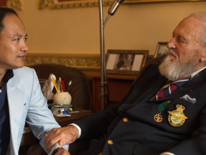 An older man sits with another man, resting his hand on the other’s arm